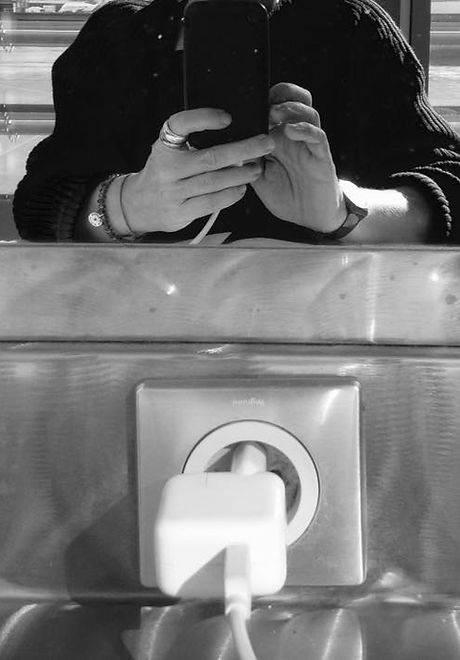 dans un miroir, des mains manipulent un smartphone. Au premier plan, une prise dans laquelle est fichée une alimentation