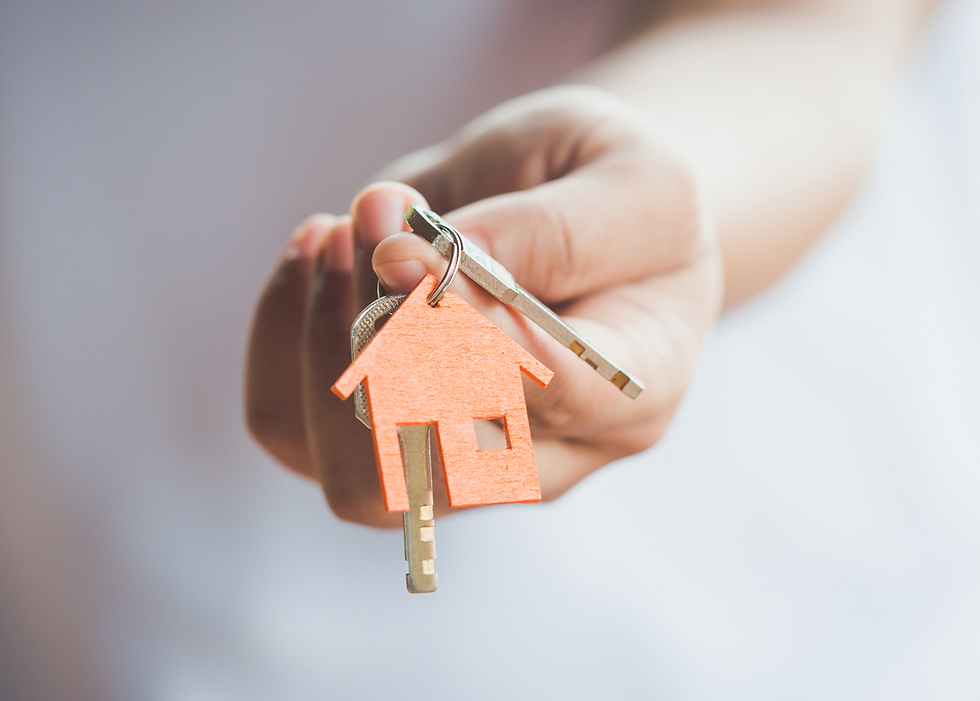Hand holding a silver key with a wooden house-shaped keychain, symbolizing real estate or home ownership, against a blurred light background.
