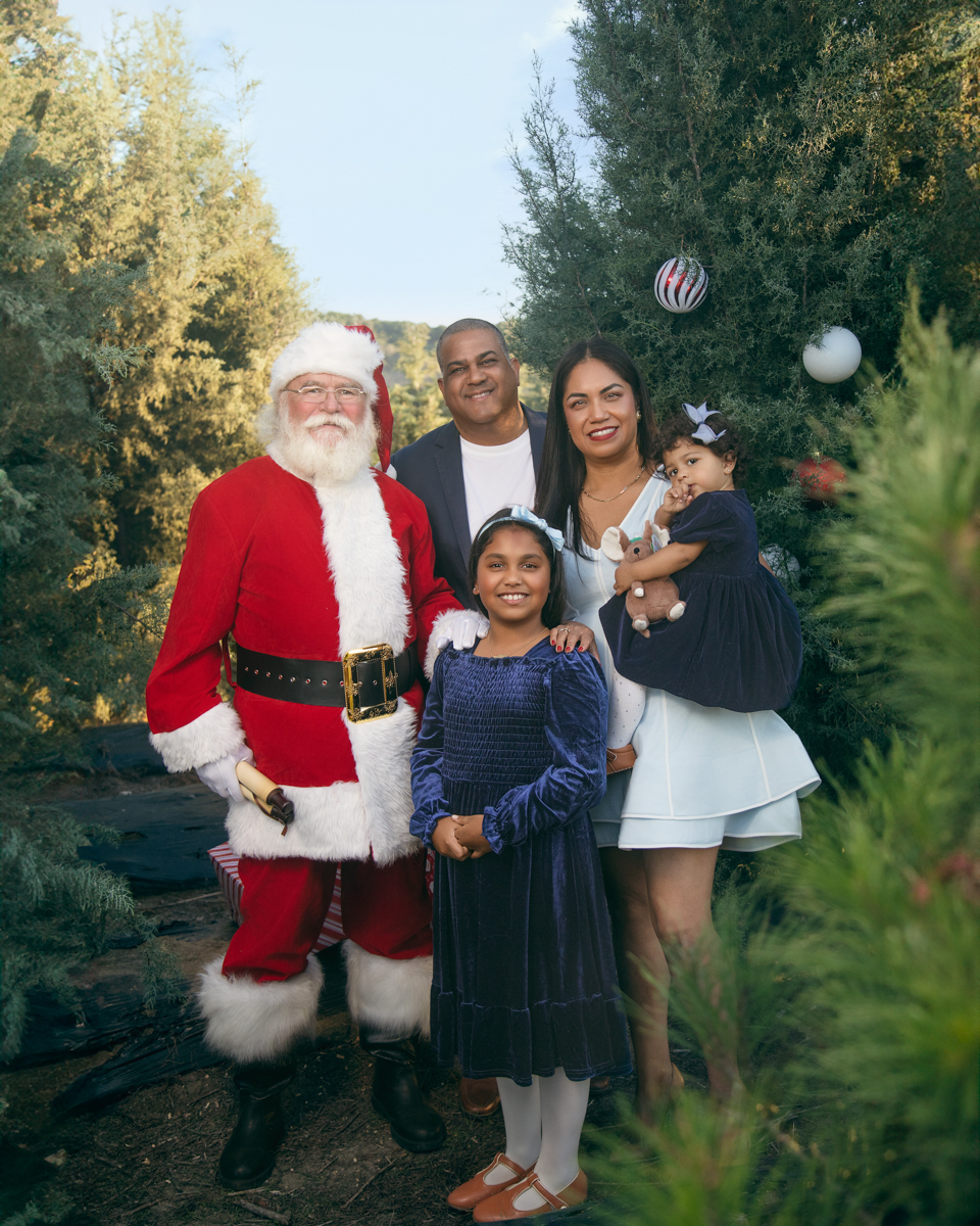 Santa poses with a family of four, mom, dad and two little girls set among fir trees at a christmas tree farm