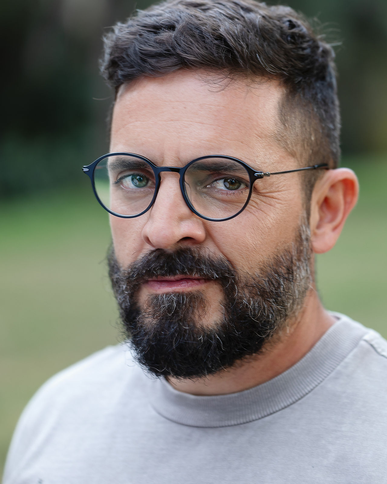 headshot of man with beard wearing glasses, a gray tee shirt at a park