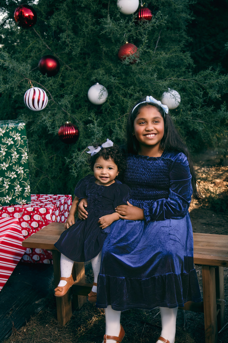 Two litlle sisters pose for sibling portraits set among fir trees at a Christmas tree farm