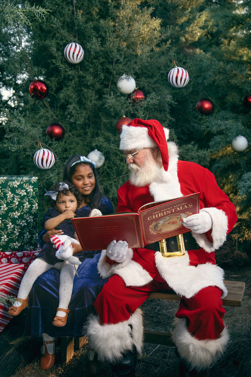 Santa reads from a big red book to two little sisters, set among fir trees decorated with chritmas ornamentss at a Christmas tree farm