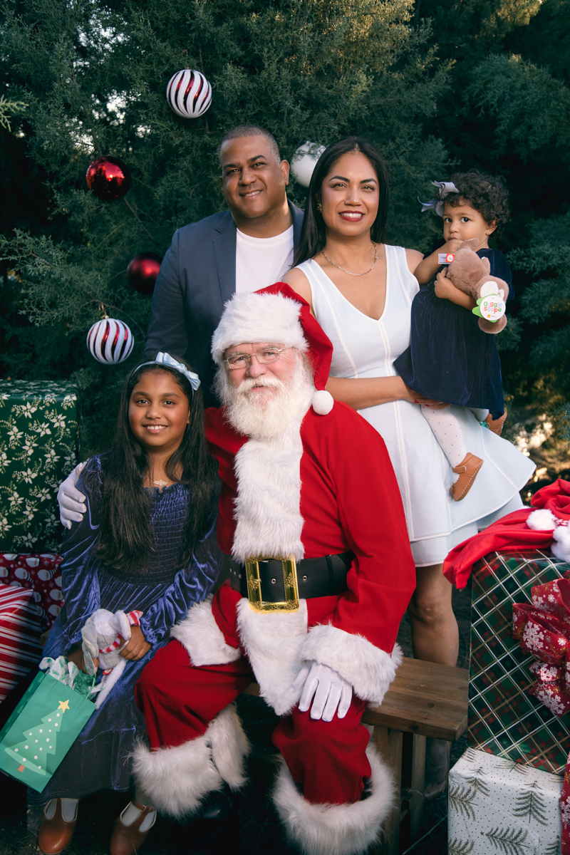 Santa poses with a family of four, mom, dad and two little girls set among decorated fir trees at a christmas tree farm