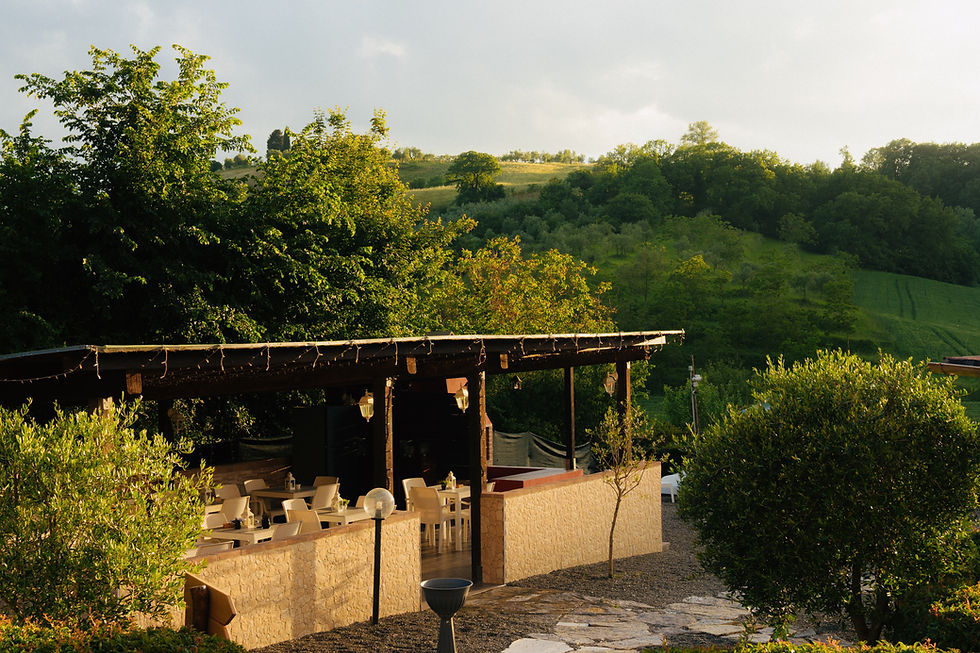 Outdoor patio with empty white tables under a wooden pergola, surrounded by lush green trees and hills in soft sunset light.