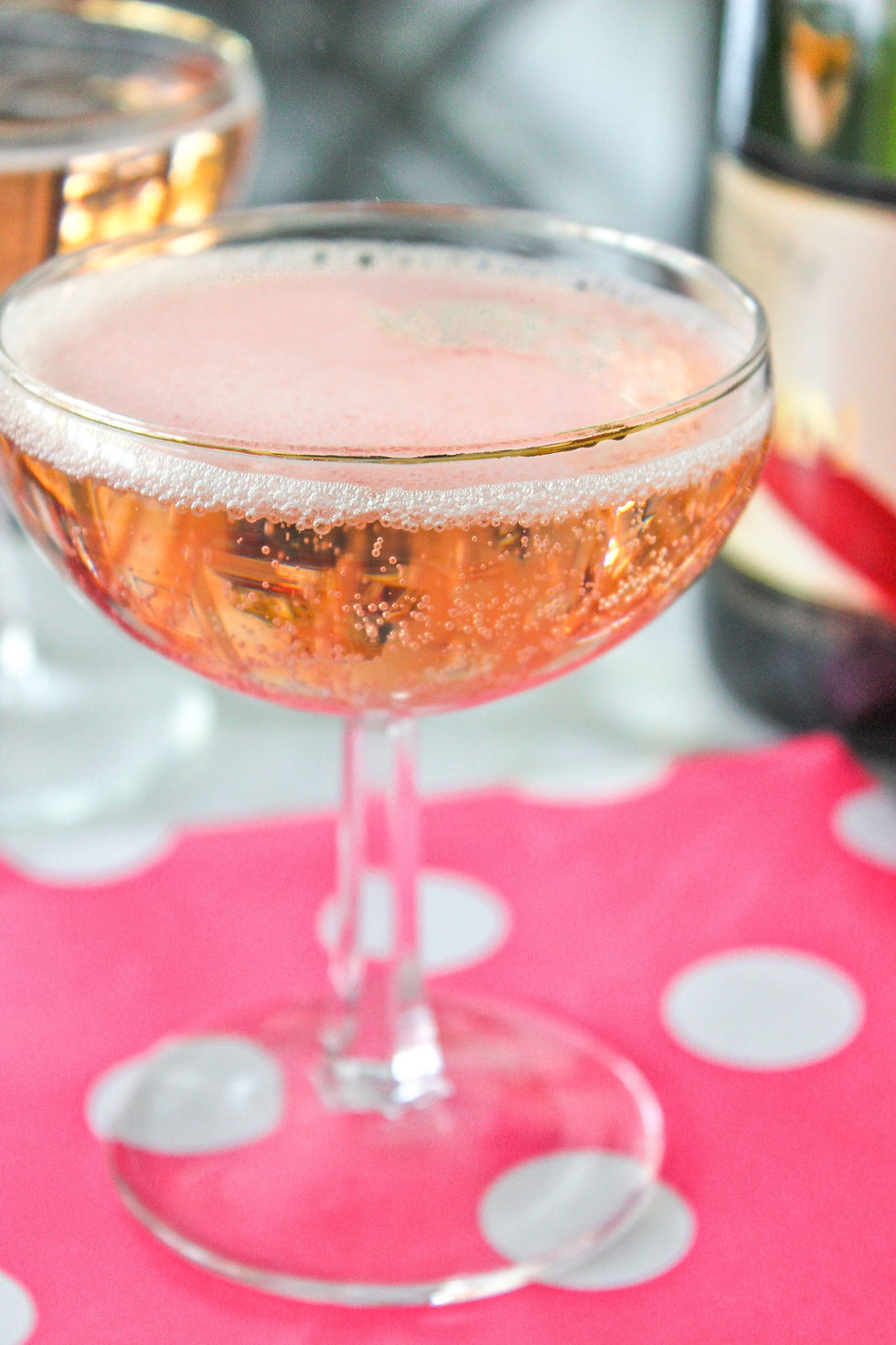 A filled champagne coupe with pink bubbly on a pink and white polka dot cloth. A bottle in the blurred background suggests celebration.