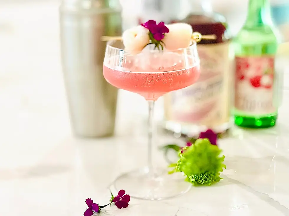 A pink cocktail in a glass with a flower and fruit garnish sits on a marble surface. Shaker and bottles in blurred background.