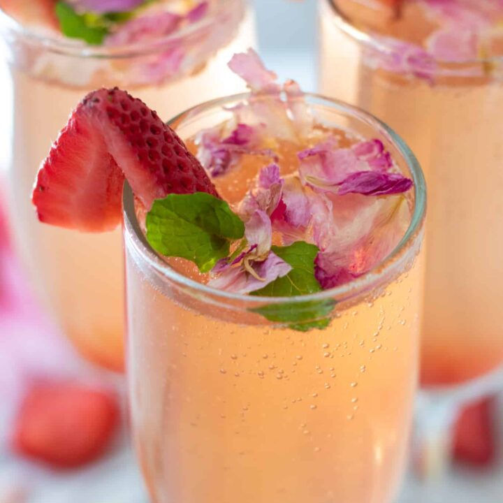 Pink sparkling drink in a glass adorned with sliced strawberry, mint leaves, and pink petals. Bright and refreshing ambiance.