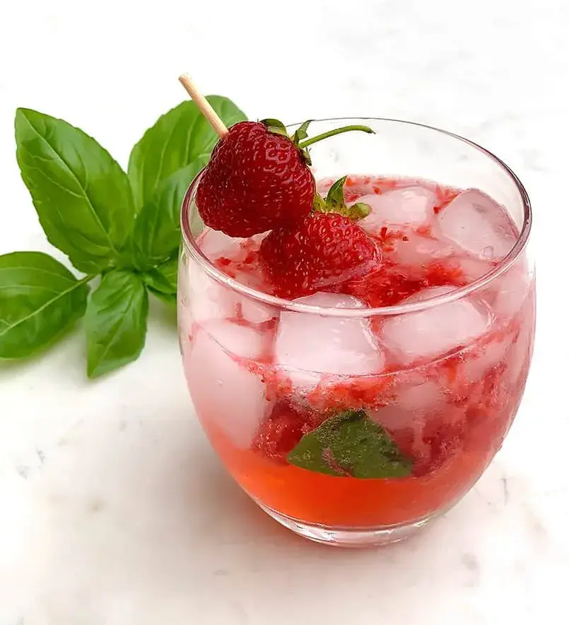 A glass of strawberry cocktail with ice, garnished with whole strawberries and basil on a white marble surface. Refreshing and vibrant.