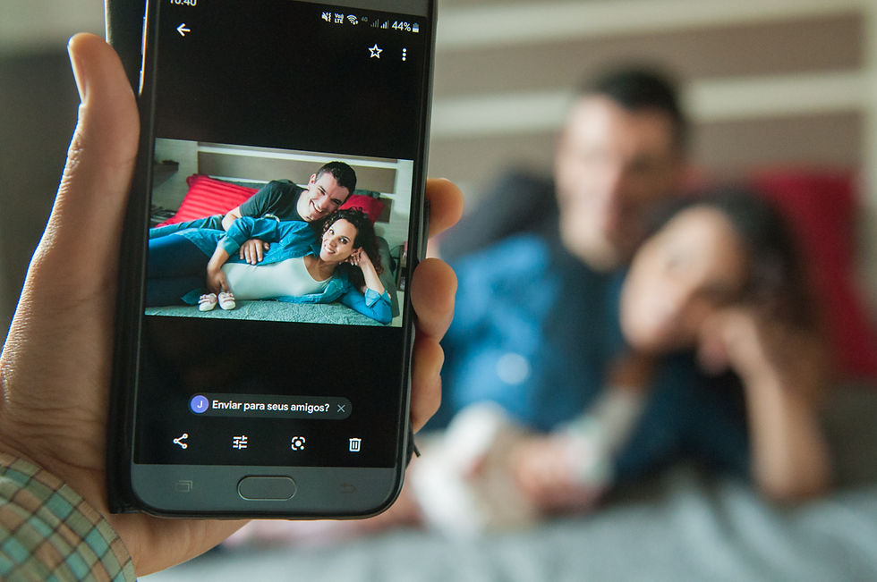 imagem à frente de um celular que mostra pai e gestante deitadas na cama com sapatinhos de bebê na frente deles, e ao fundo a família desfocada