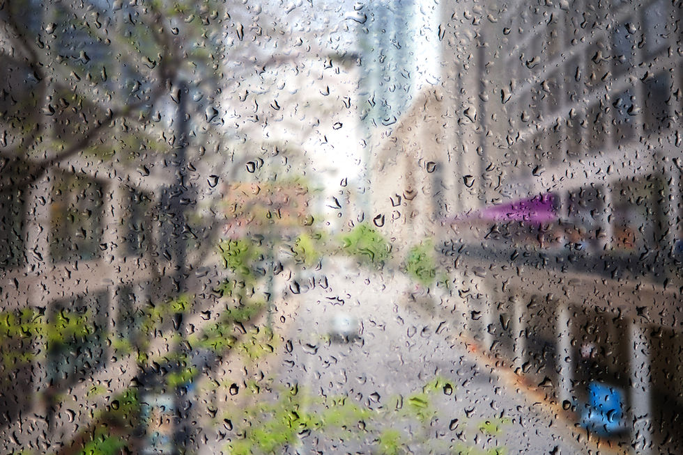Blurred view of a city street seen through a rain-streaked window, with buildings and trees softened by water on the glass, suggesting a quiet moment of reflection during ongoing movement.