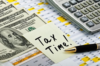 A sticky note reading Tax Time sits atop paperwork near a pen, cash and a calculator