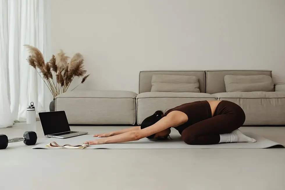 Woman with fibromyalgia in child’s pose on a yoga mat, dressed in brown activewear. A nearby laptop and hand weights suggest a guided home workout for fibromyalgia symptom relief in a cozy, neutral-toned living room.