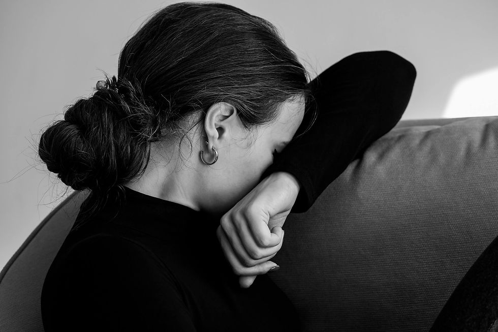 Black and white image of a woman with fibromyalgia wearing a black shirt, leaning her face on her arm while seated on a sofa. Her distressed expression reflects the emotional and physical toll of chronic fibromyalgia pain in a neutral-toned room.