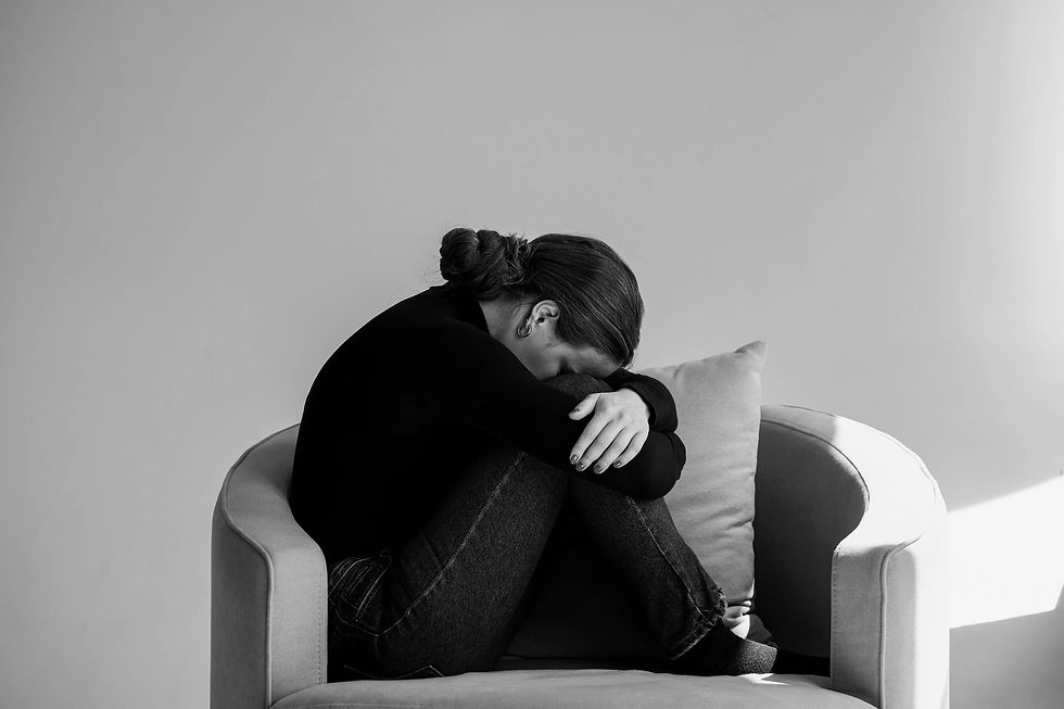 Woman with fibromyalgia wearing a black sweater and jeans sits on a chair, hugging her knees with her head down. The minimalistic room emphasizes the emotional weight and isolation often experienced with chronic pain and fatigue.