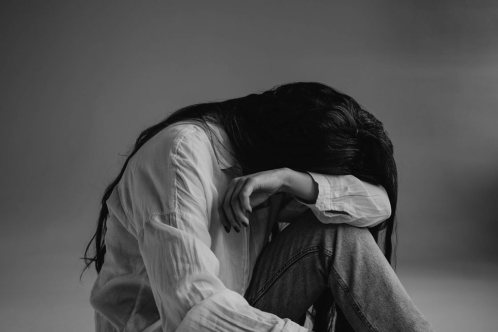 Woman with fibromyalgia in a white shirt and jeans sits curled up with her head on her knees against a gray background. Black and white image expresses emotional exhaustion, chronic pain, and isolation.