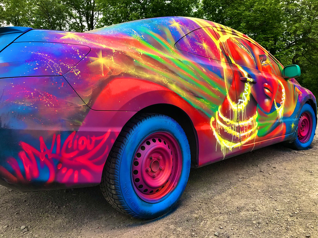 Voiture peinte par MALICIOUZ au Makadam Jam 2021, montrant un visage féminin majestueux et des motifs lumineux sur une carrosserie colorée dans des couleurs bleues, rouge, rose fluo et autres couleurs vibrantes