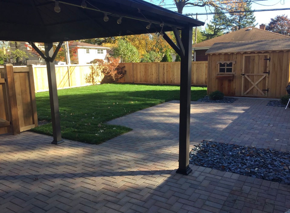 A wide view of a completed residential backyard featuring a large Unilock paver patio in a herringbone pattern, and a premium green sod.