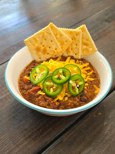 a bowl of chili on a dark wood table. The chili is garnished with cheese, fresh jalapenos, and saltine crackers have been place around the back rim of the bowl.