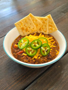 a bowl of chili on a dark wood table. The chili is garnished with cheese, fresh jalapenos, and saltine crackers have been place around the back rim of the bowl.