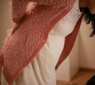 Pregnant woman wearing a white lace dress with a fuzzy salmon-colored cardigan bent over in pain with her hands on her lower back