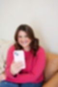 Woman smiling, taking a selfie with a smartphone. She wears a pink sweater, sitting on a beige couch. Relaxed mood, neutral wall background.branding session with Makana Photography ins Southern Pines and Aberdeen