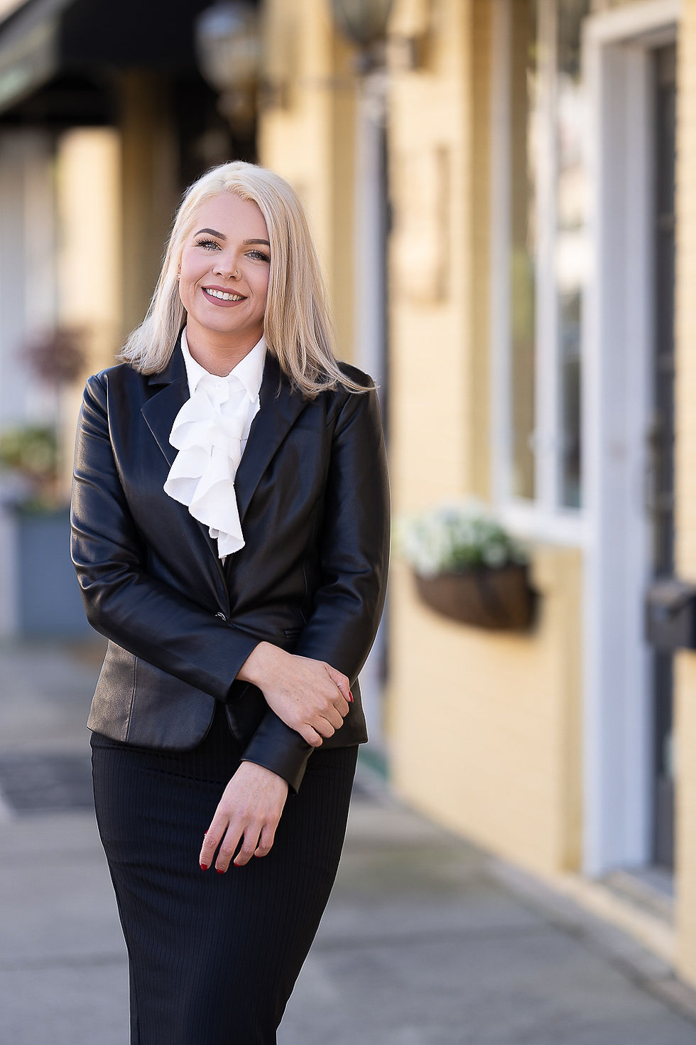 Smiling woman with blonde hair in a black leather jacket and white blouse, standing on a sunny street with yellow building background.Downtown Aberdeen NC in her realtor headshot and branding session by Makana photography, a commercial moore county photo studio