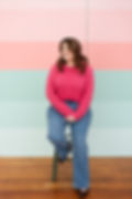 Woman in pink sweater and blue jeans sits on a stool, smiling, against a colorful striped pastel wall. Warm, cheerful mood.branding session with Makana Photography ins Southern Pines and Aberdeen