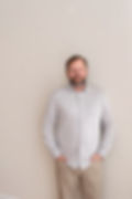 Man with a beard and hands in pockets stands smiling against a plain beige wall, wearing a light gray shirt and khaki pants. Calm mood.Branding session in Southern Pines, Aberdeen, pinehurst in Moore county