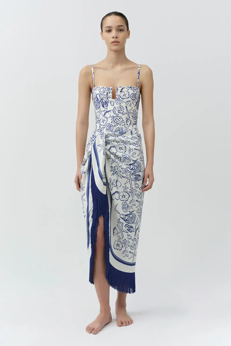 Barefoot model in a white-and-blue seashell-print dress with fringe stands against a plain white studio background.