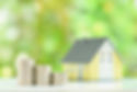 Stacks of coins increase in size, leading to a small yellow house in a green, blurred background, suggesting financial growth.