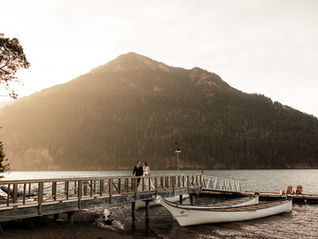 Photo of a bride and groom walking up a dock on Lake Crescent at NatureBridge in Olympic National Park. There are two canoes in the water and two chairs at the end of the dock. The sun is setting behind a mountain and the light is warm and sunny in the photo. 