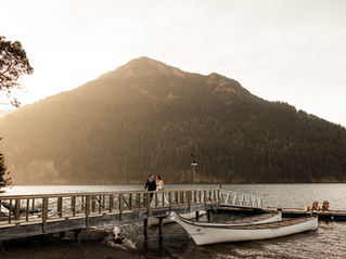 Photo of a bride and groom walking up a dock on Lake Crescent at NatureBridge in Olympic National Park. There are two canoes in the water and two chairs at the end of the dock. The sun is setting behind a mountain and the light is warm and sunny in the photo. 
