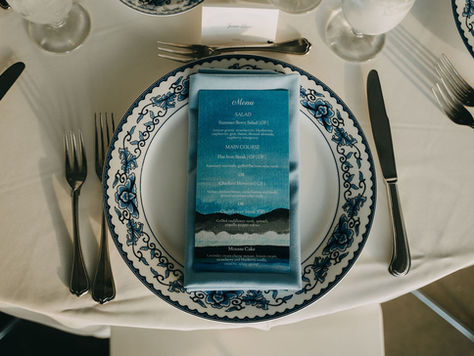 Top-down photo of a place setting at a dinner table with white tablecloth, silver flatware around the plate, a white plate with a blue floral pattern around the rim, a folded light blue napkin on top of the plate, and a dinner menu with an illustrated ocean/mountain/sky view.