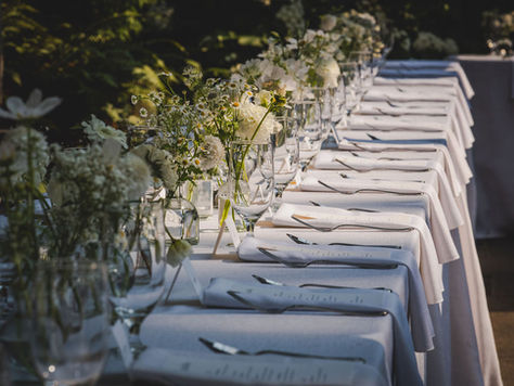 Photo of a long dinner table in sunlight with white linens, silverware, and menus set. The middle of the table has an assortment of white and green florals in vases.