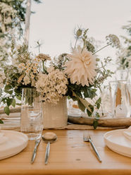Close up photo of a wedding reception dinner table. The table is a light wood. The plates and napkins are white with silverware and simple clear water glasses next to the plates. There is a centerpiece in a white vase with assorted white flowers inside.
