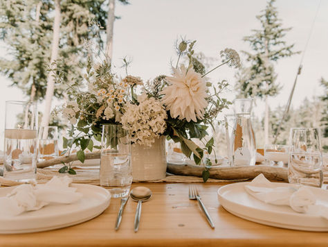 Close up photo of a wedding reception dinner table. The table is a light wood. The plates and napkins are white with silverware and simple clear water glasses next to the plates. There is a centerpiece in a white vase with assorted white flowers inside.