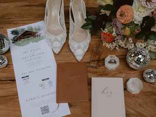 Flat lay of disco wedding details featuring elegant stationery suite, rings, disco balls, wildflower bridal bouquet perfume, and delicate styling elements arranged on a wood backdrop in natural light at the Evergreen Gardens