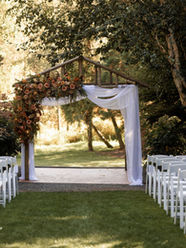 Photo of a wedding ceremony with rows of white chairs facing a wooden A-frame altar that is decorated with a large floral installation of pink and orange flowers on one side with white drapery on the other side. There are large trees in the background with bright sunlight hitting the trees.