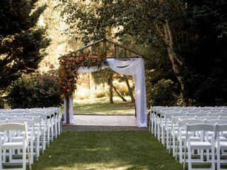 Photo of a wedding ceremony with rows of white chairs facing a wooden A-frame altar that is decorated with a large floral installation of pink and orange flowers on one side with white drapery on the other side. There are large trees in the background with bright sunlight hitting the trees.