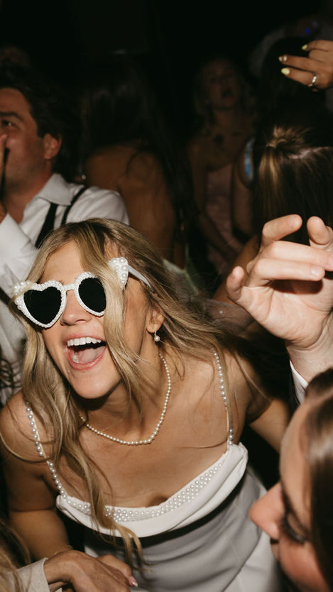 Photo of a bride and groom dancing with their guests at a wedding reception. The bride is wearing white, heart-shaped sun galsses and the groom's shirt is partially unbuttoned. They are singing and smiling surrounded by guests that are dancing with their hands in the air.
