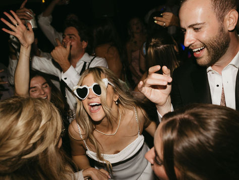 Photo of a bride and groom dancing with their guests at a wedding reception. The bride is wearing white, heart-shaped sun galsses and the groom's shirt is partially unbuttoned. They are singing and smiling surrounded by guests that are dancing with their hands in the air.