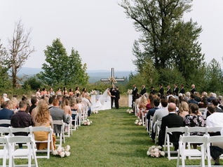 Photo of a wedding ceremony in progress. The bride and groom are holding hands at the altar in front of a large black and white cross. Their guests are seated in white chairs watching the ceremony. They have a large wedding party standing up with them. The aisles are lined on either side with white roses.