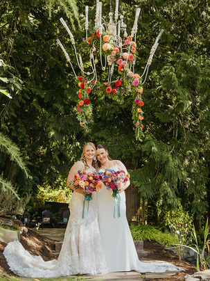 Photo of two brides on their wedding day. They are leaning into each other holding their bouquets. They are standing below a huge white outdoor chandelier that is decorated with cascading flowers. They are surrounded by lush trees in bright sunlight. 