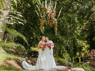 Photo of two brides on their wedding day. They are leaning into each other holding their bouquets. They are standing below a huge white outdoor chandelier that is decorated with cascading flowers. They are surrounded by lush trees in bright sunlight.