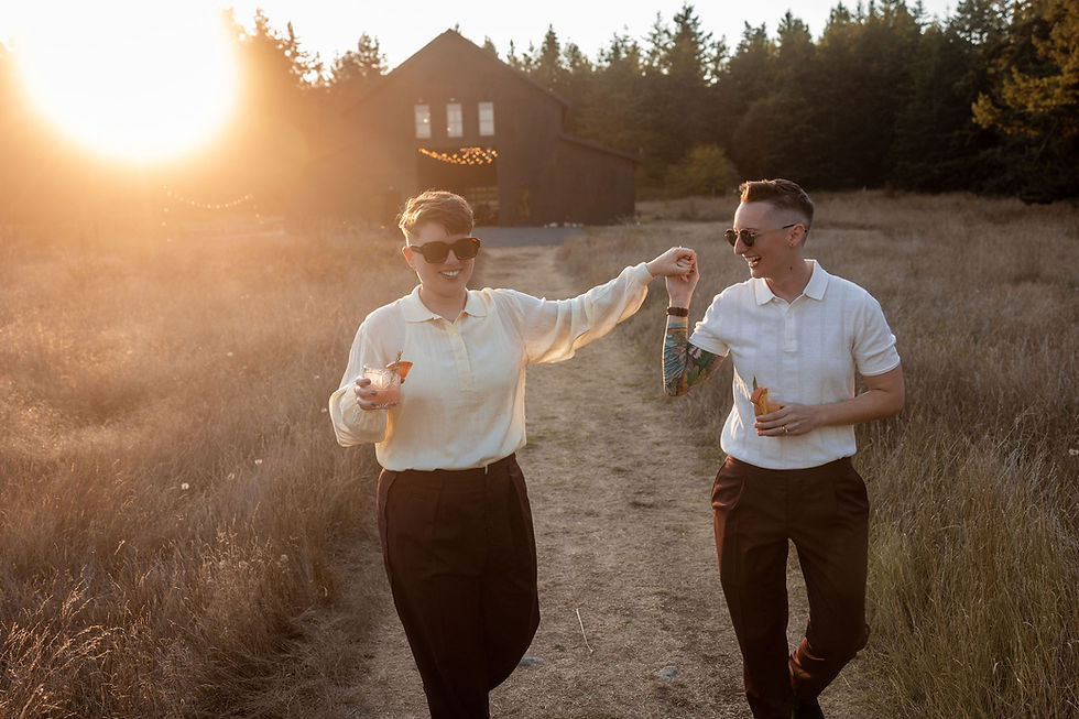 Photo of a queer couple at their wedding. They are holding hands and carrying cocktails at golden hour walking away from their reception space with the sun setting behind them. They are smiling and laughing together.