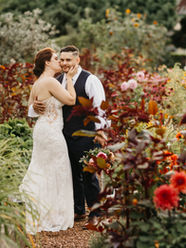 A couple on their wedding day standing in a garden. They are surrounded by lush green plants with pink, orange, and yellow flowers. They are hugging and the bridge is kissing the groom on the cheek while the groom looks at the camera.