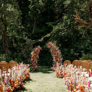 Photo of a wedding ceremony looking down the aisle at the altar which is framed by a large arch covered entirely in brightly colored flowers. There are large floral installments lining both sides of the aisle with tan chairs facing forward. Behind the altar there are large, dark green trees.