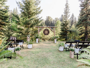 Photo of a wedding ceremony location on a grass lawn with large trees in the distance. There are black folding chairs set up in rows with plants lining the aisle. There is a large wooden arbor with a circle art piece hanging from the center with a celtic design.