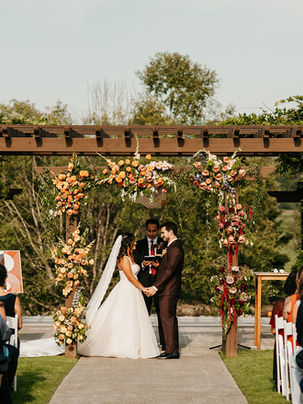 Photo of an outdoor wedding ceremony in progress with bride and groom holding hands in front of an officiant and their guests sitting in chairs. They are framed by an arbor covered in pink, yellow, and orange flowers. There are trees and grass in the background. 