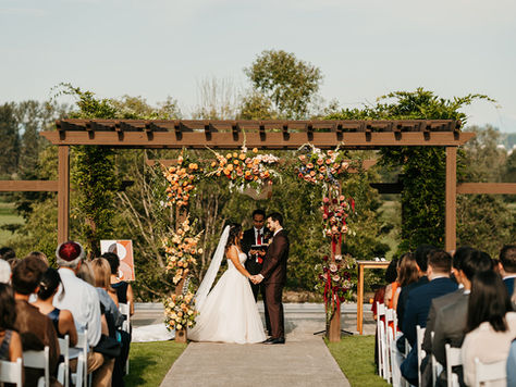 Photo of an outdoor wedding ceremony in progress with bride and groom holding hands in front of an officiant and their guests sitting in chairs. They are framed by an arbor covered in pink, yellow, and orange flowers. There are trees and grass in the background.